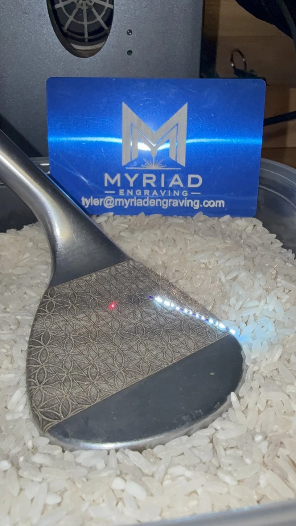 Steel golf club head being laser engraved with a geometric flower-of-life pattern, resting in a bed of rice with a blue Myriad Engraving card standing behind it
