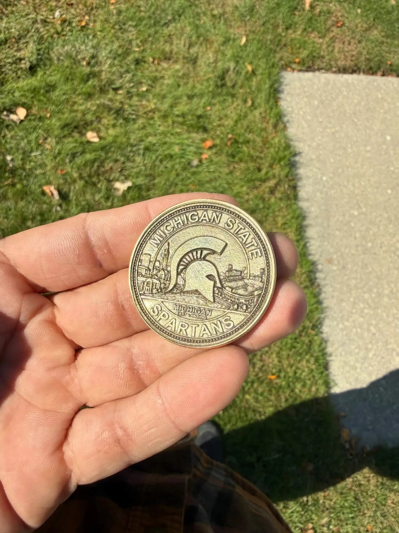 Antique brass Michigan State Spartans challenge coin engraved with a Spartan helmet, campus landmarks, and beaded borders, held in an open hand